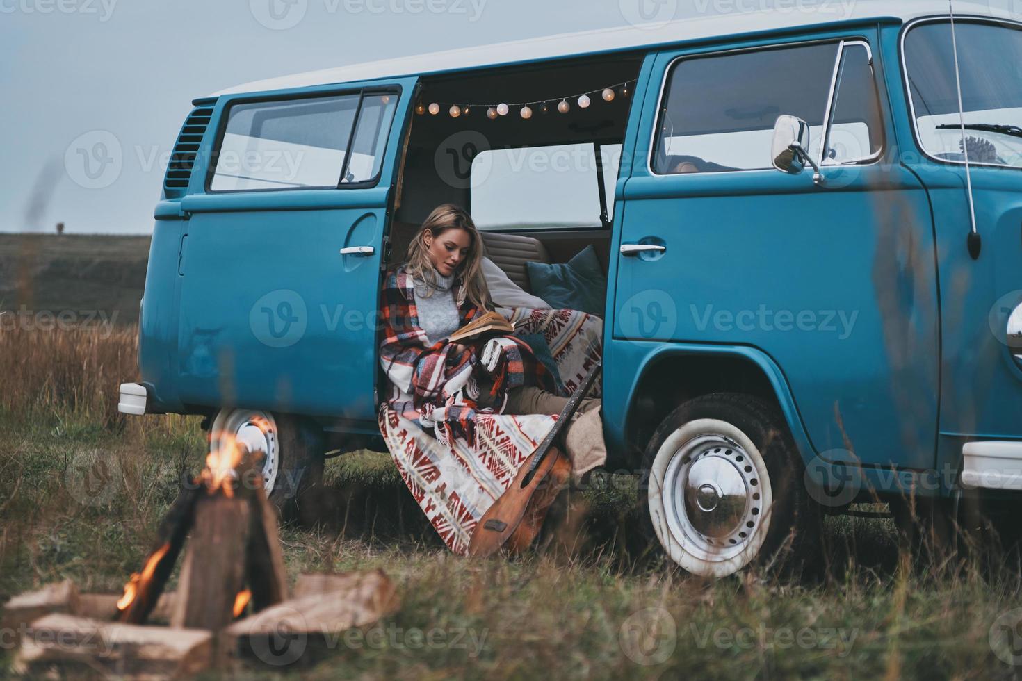 genießt ihr Lieblingsbuch. Attraktive junge Frau, die mit einer Decke bedeckt ist und ein Buch liest, während sie im Inneren des blauen Minivans im Retro-Stil sitzt foto