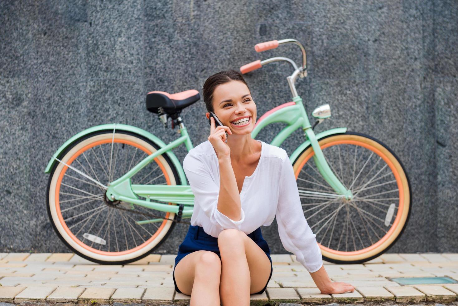 gutes Gespräch mit Freund. Attraktive junge lächelnde Frau, die mit dem Handy spricht, während sie am Straßenrand in der Nähe ihres Oldtimer-Fahrrads sitzt foto