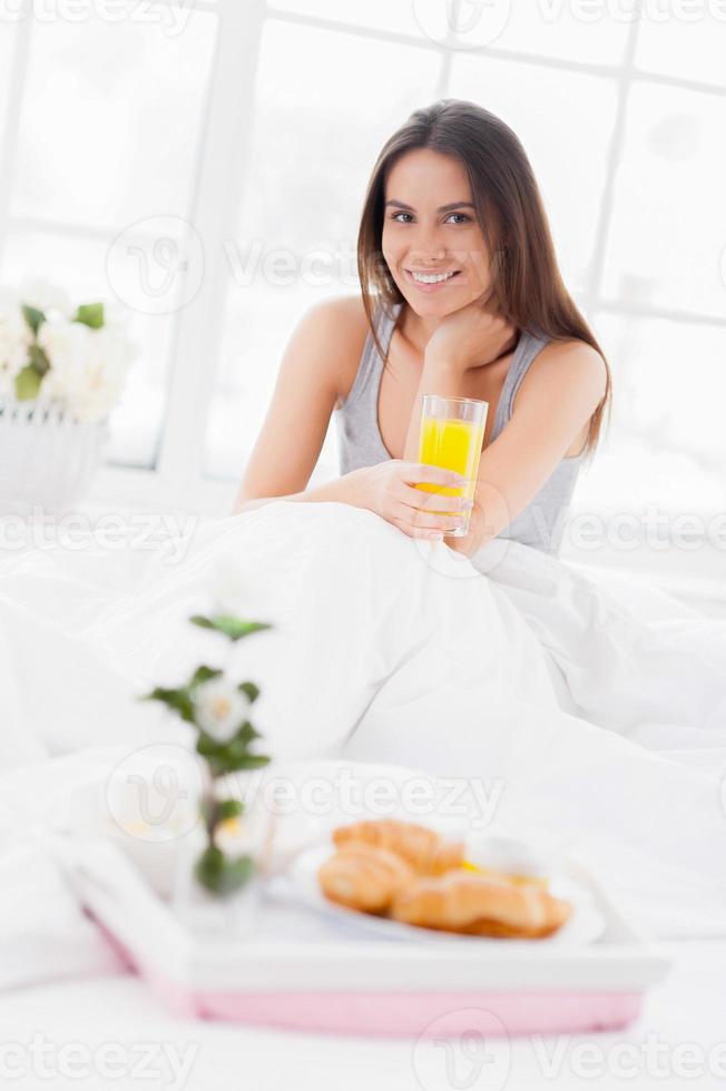 Gesunder Start in den Tag. fröhliche junge lächelnde frau, die ein glas mit saft hält, während sie mit einem frühstück auf einem tablett im bett sitzt foto