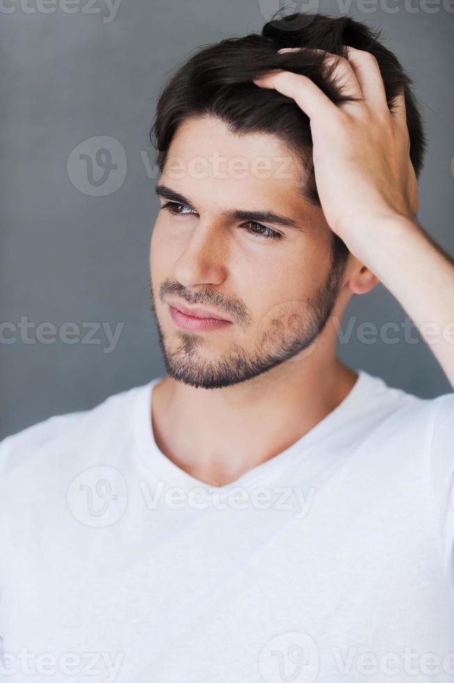 sah früher gut aus. hübscher junger Mann, der die Hand im Haar hält und wegschaut, während er vor grauem Hintergrund steht foto