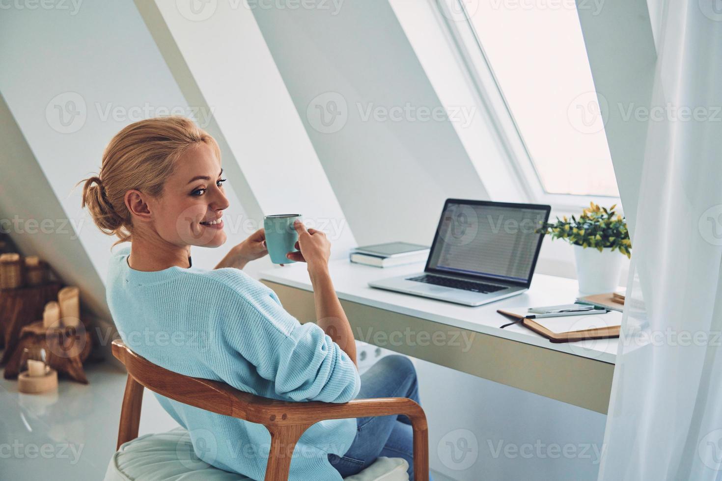 Rückansicht einer jungen lächelnden Frau, die ein heißes Getränk genießt, während sie zu Hause arbeitet foto