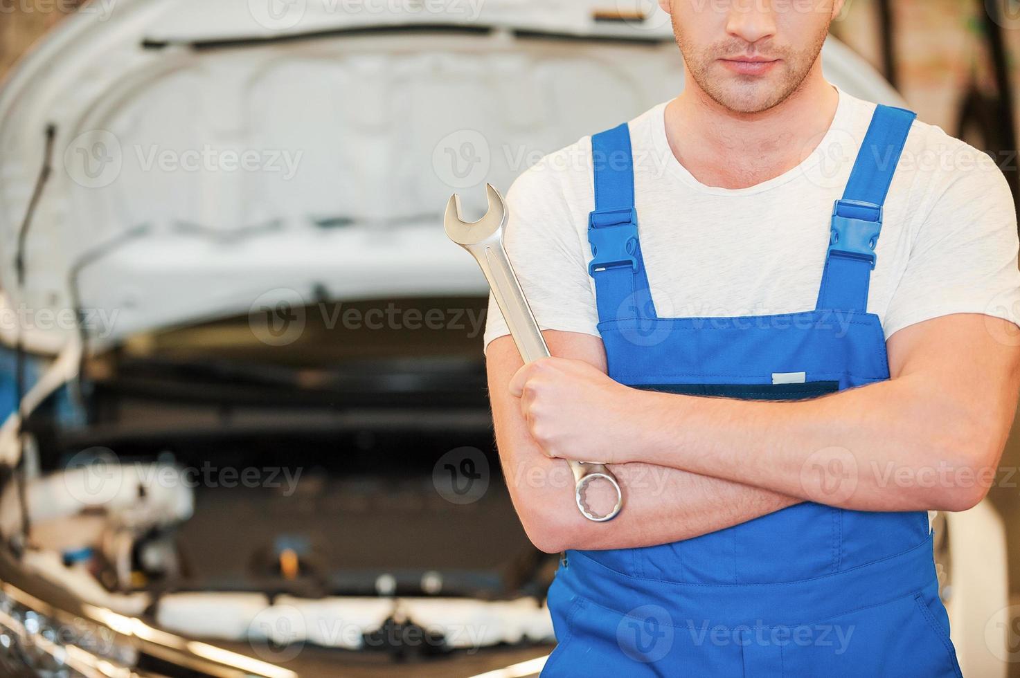 bereit zu arbeiten. Nahaufnahme eines selbstbewussten jungen Mannes, der einen Schraubenschlüssel hält, während er in der Werkstatt mit dem Auto im Hintergrund steht foto