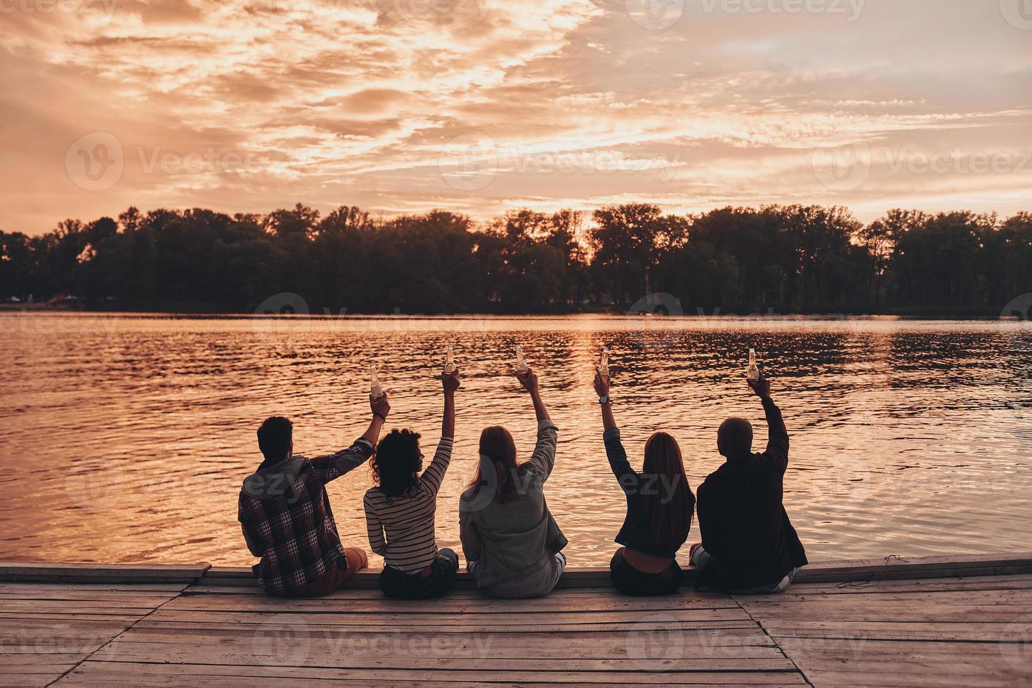 schönen Sonnenuntergang genießen. Rückansicht junger Menschen in Freizeitkleidung, die mit einer Bierflasche anstoßen, während sie auf dem Pier sitzen foto