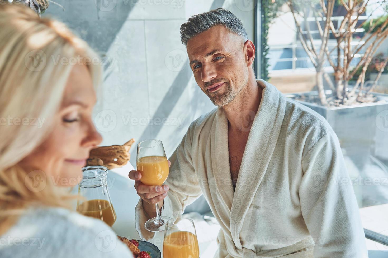 Schönes reifes Paar in Bademänteln, das zusammen frühstückt, während es Zeit in der heimischen Küche verbringt foto