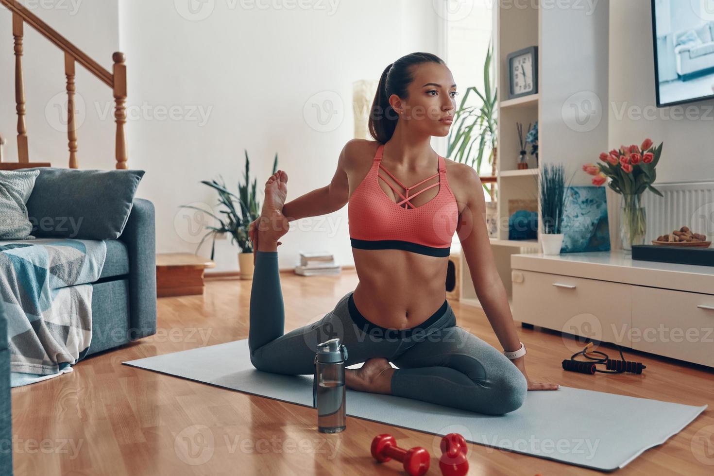 Schöne junge Frau in Sportkleidung, die sich auf der Trainingsmatte ausdehnt, während sie Zeit zu Hause verbringt foto