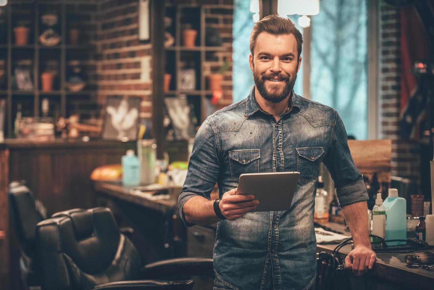 Mit digitalen Technologien das Geschäft am Laufen halten. Fröhlicher junger bärtiger Mann, der in die Kamera schaut und ein digitales Tablet hält, während er im Friseursalon steht foto