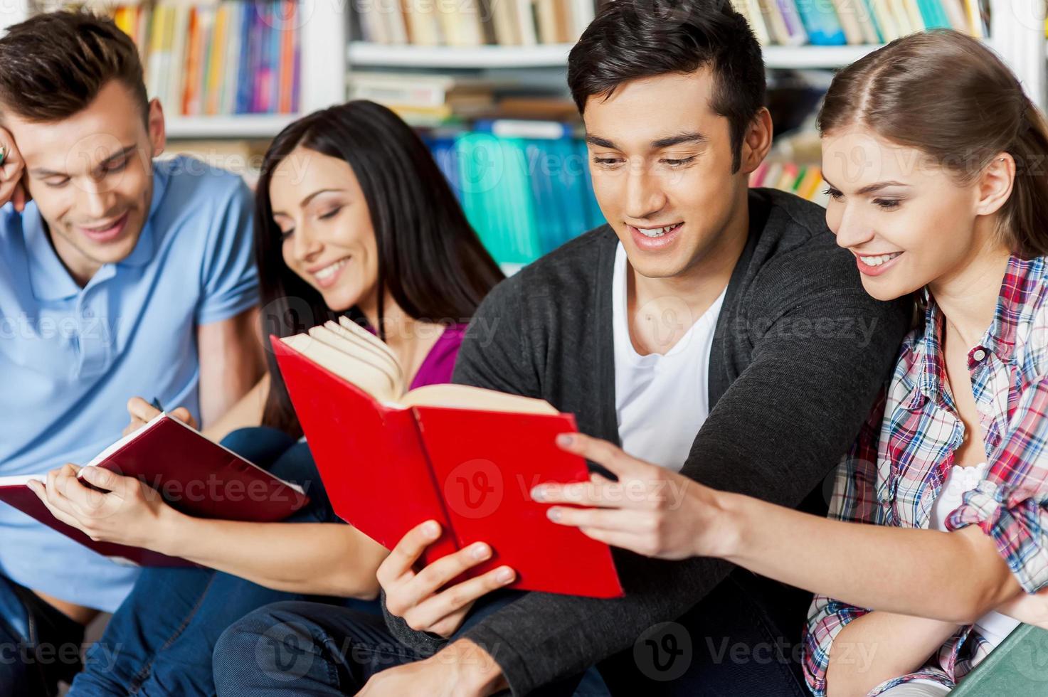 Studenten in der Bibliothek. Vier fröhliche Studenten, die zusammen ein Buch lesen, während sie in einer Bibliothek gegen ein Bücherregal sitzen foto