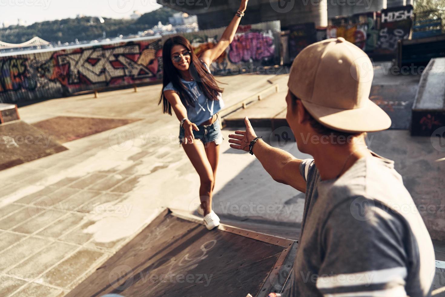 Schöne junge Frau, die versucht, zu ihrer Freundin aufzusteigen, während sie Zeit im Skatepark im Freien verbringt foto