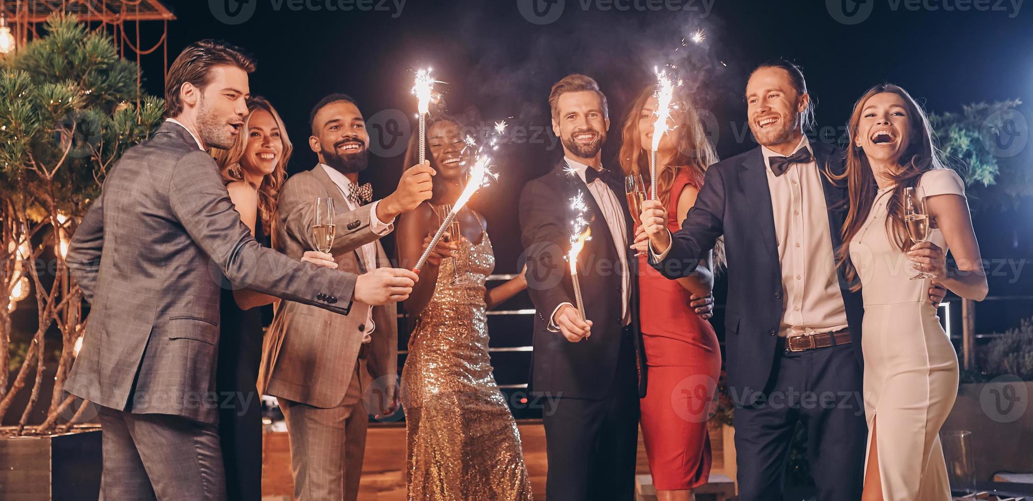 Gruppe schöner Menschen in formeller Kleidung, die Wunderkerzen halten und lächeln, während sie Zeit auf einer Party im Freien verbringen foto