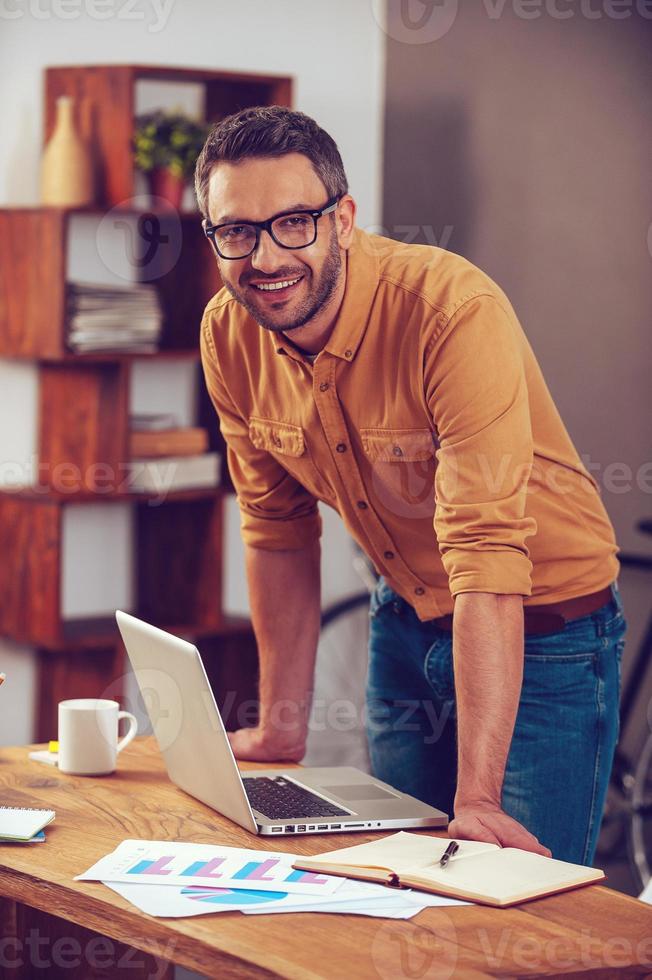 in jeder Situation positiv bleiben. hübscher junger Mann, der in die Kamera schaut und lächelt, während er in der Nähe seines Arbeitsplatzes im Büro steht foto