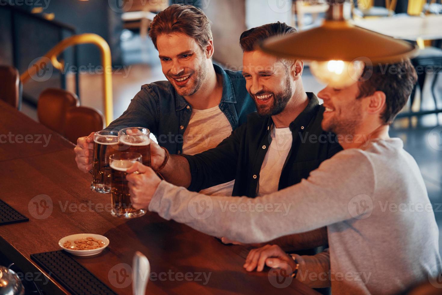 Blick von oben auf glückliche junge Männer in Freizeitkleidung, die sich gegenseitig mit Bier anstoßen und lächeln, während sie an der Bartheke im Pub sitzen foto