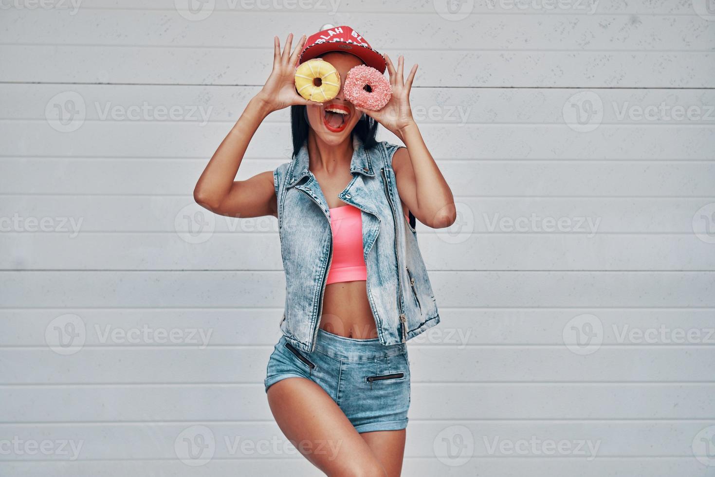 Sie weiß nicht, was die Langeweile ist. Verspielte junge Frauen, die Donuts gegen ihre Augen halten und lächeln, während sie gegen das Garagentor stehen foto