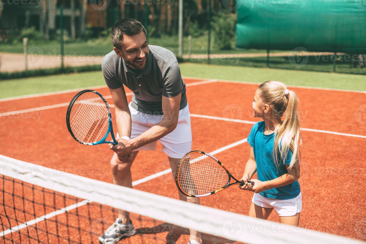 Tennis üben. Fröhlicher Vater in Sportkleidung bringt seiner Tochter das Tennisspielen bei, während beide auf dem Tennisplatz stehen foto