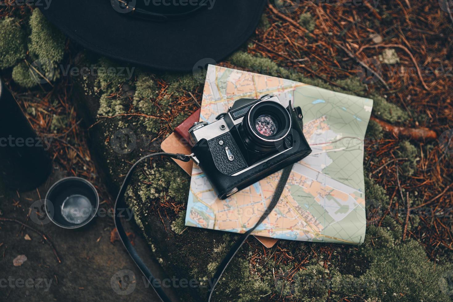 Dinge, die man im Wald braucht. Nahaufnahme von oben auf Digitalkamera, Karte, Hut und isolierten Getränkebehälter auf dem Boden im Wald foto