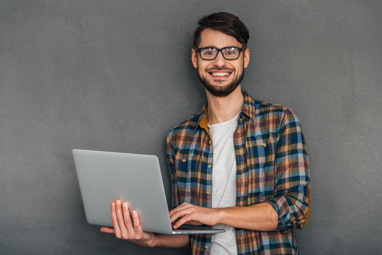 bereit, Ihnen zu helfen. Selbstbewusster junger Mann, der seinen Laptop hält und mit einem Lächeln in die Kamera schaut, während er vor grauem Hintergrund steht foto