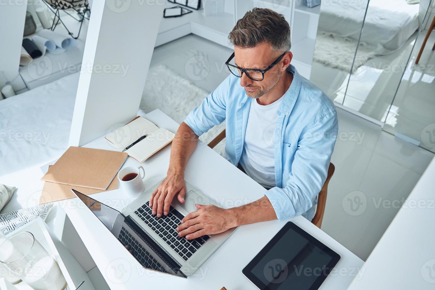 Draufsicht eines selbstbewussten reifen Mannes, der am Laptop arbeitet, während er am Schreibtisch sitzt foto