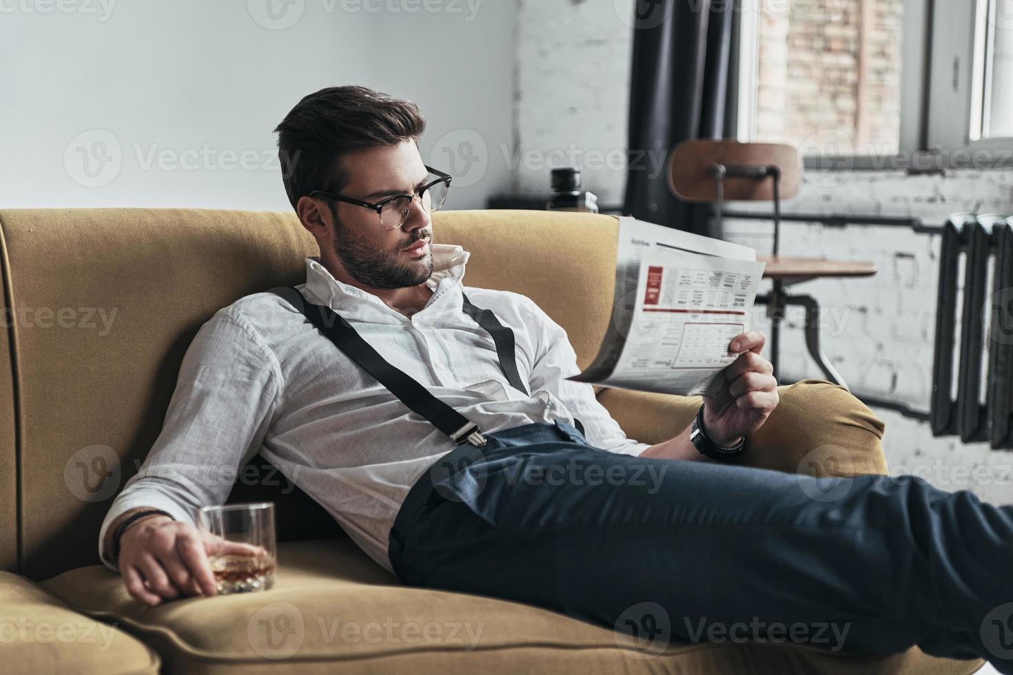 interessanter Artikel. Stilvoll gekleideter junger Mann, der eine Zeitung liest und ein Glas hält, während er auf dem Sofa sitzt foto
