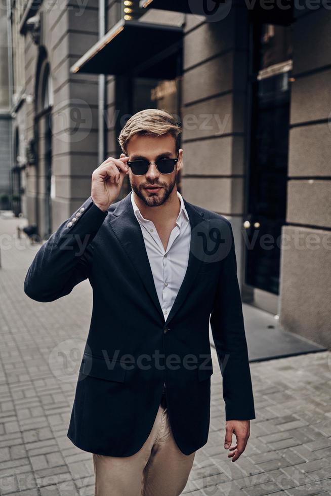 in seinem eigenen Stil. hübscher junger Mann in eleganter Freizeitkleidung, der seine Brille anpasst, während er durch die Stadtstraße geht foto