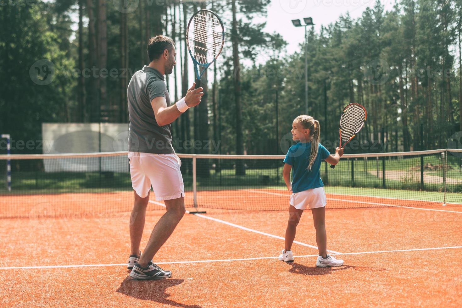 gemeinsam trainieren. Rückansicht des Vaters in voller Länge in Sportkleidung, die seiner Tochter das Tennisspielen beibringt, während beide auf dem Tennisplatz stehen foto