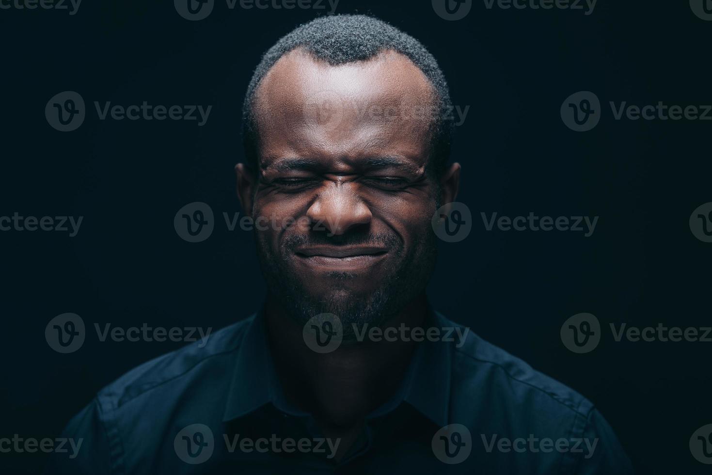 verzweifelt fühlen. Porträt eines jungen afrikanischen Mannes, der ein Gesicht macht, während er die Augen geschlossen hält und vor schwarzem Hintergrund steht foto