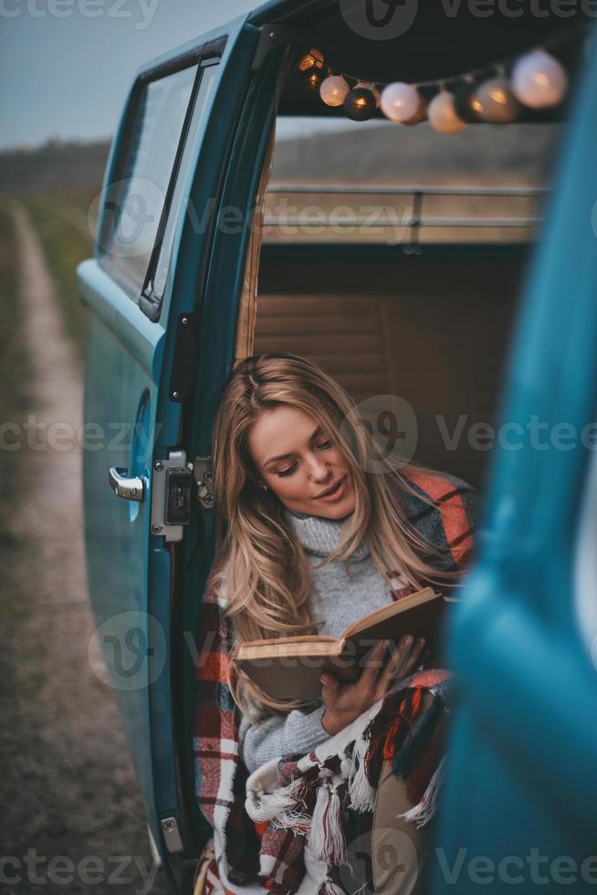 am Lesen beteiligt. Attraktive junge Frau, die mit einer Decke bedeckt ist und ein Buch liest, während sie im Inneren des blauen Minivans im Retro-Stil sitzt foto