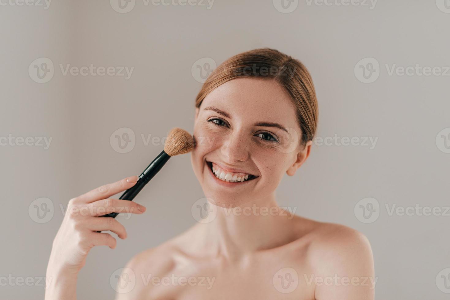 Zeit für sich. Schöne junge Frau mit Sommersprossen im Gesicht, die einen Make-up-Pinsel hält und in die Kamera schaut, während sie vor dem Hintergrund steht foto