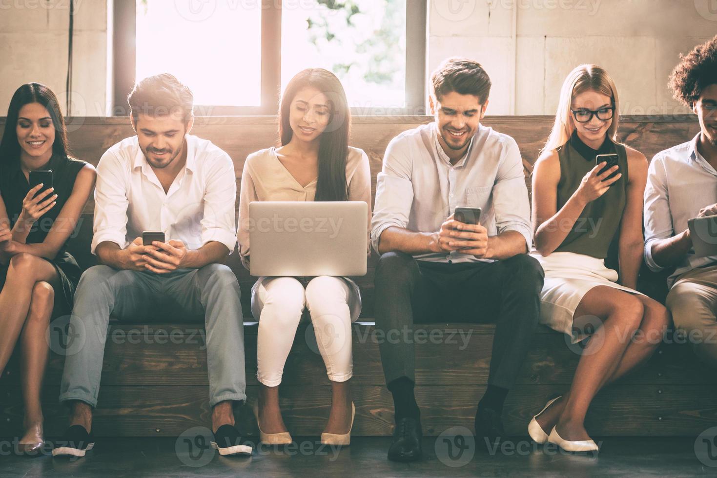 digitales Leben. Vorderansicht junger Menschen, die verschiedene Gadgets verwenden, während sie nahe beieinander in einer Reihe sitzen foto