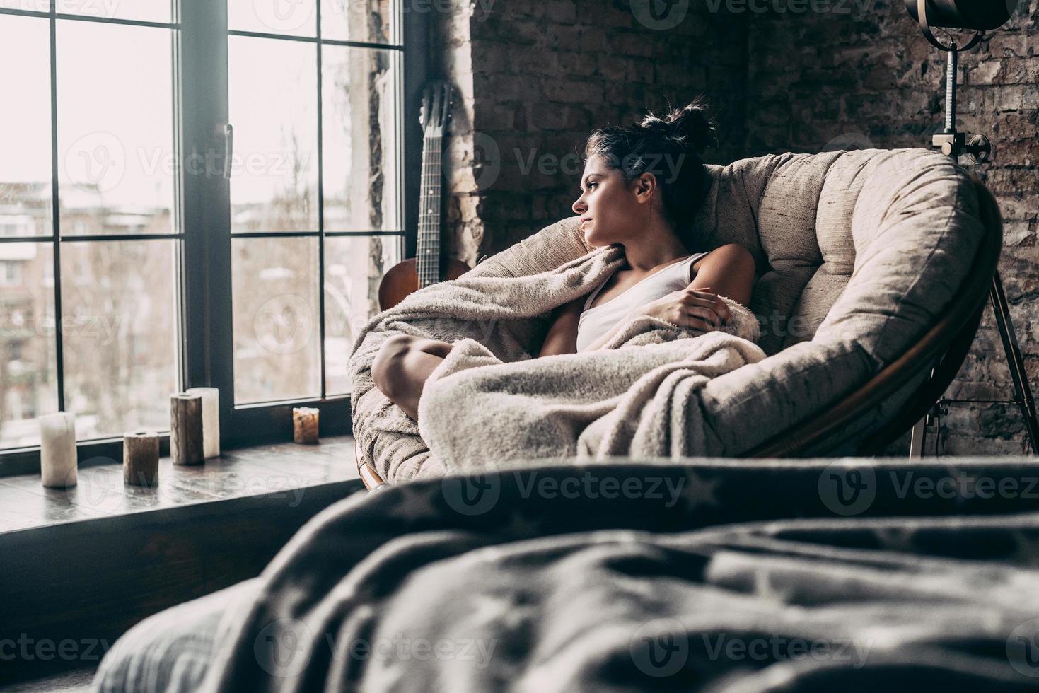 fauler tag zu hause. Schöne junge Frau, die mit einer Decke bedeckt ist und durch das Fenster schaut, während sie zu Hause in einem großen bequemen Stuhl sitzt foto