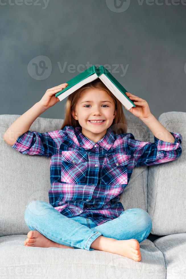 Lassen Sie uns Verstecken spielen und ein fröhliches kleines Mädchen suchen, das ein Buch über dem Kopf hält und mit einem Lächeln in die Kamera schaut, während es zu Hause in Lotusposition auf der Couch sitzt foto