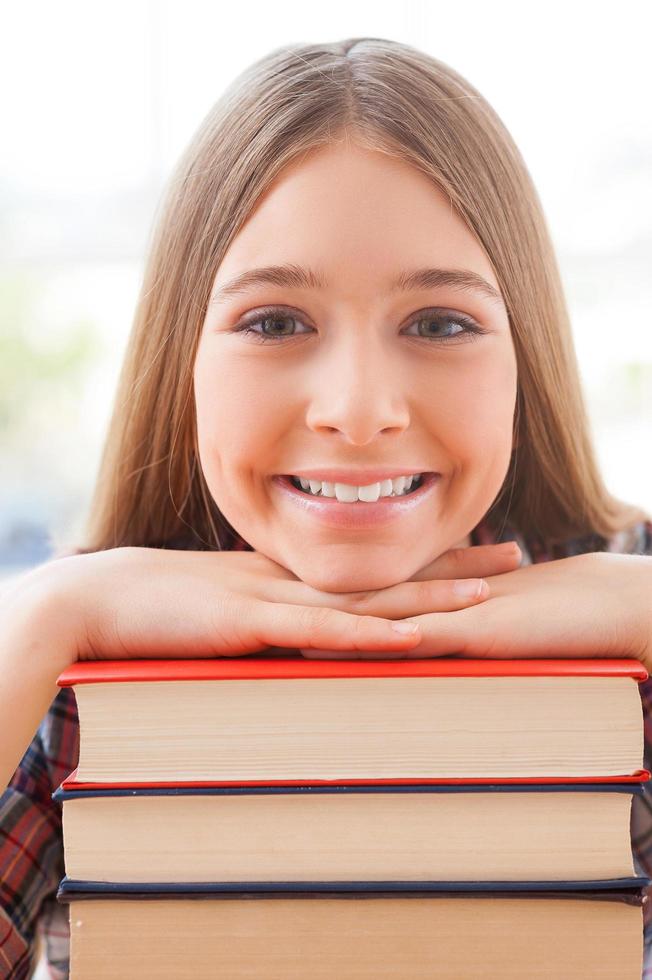 klug und selbstbewusst. Fröhliches Teenager-Mädchen, das sich an den Bücherstapel lehnt und lächelt foto