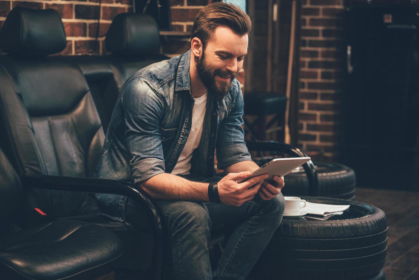 auf Termin warten. hübscher junger bärtiger mann, der mit einem lächeln auf einem digitalen tablet arbeitet, während er im friseursalon in einem bequemen stuhl sitzt foto
