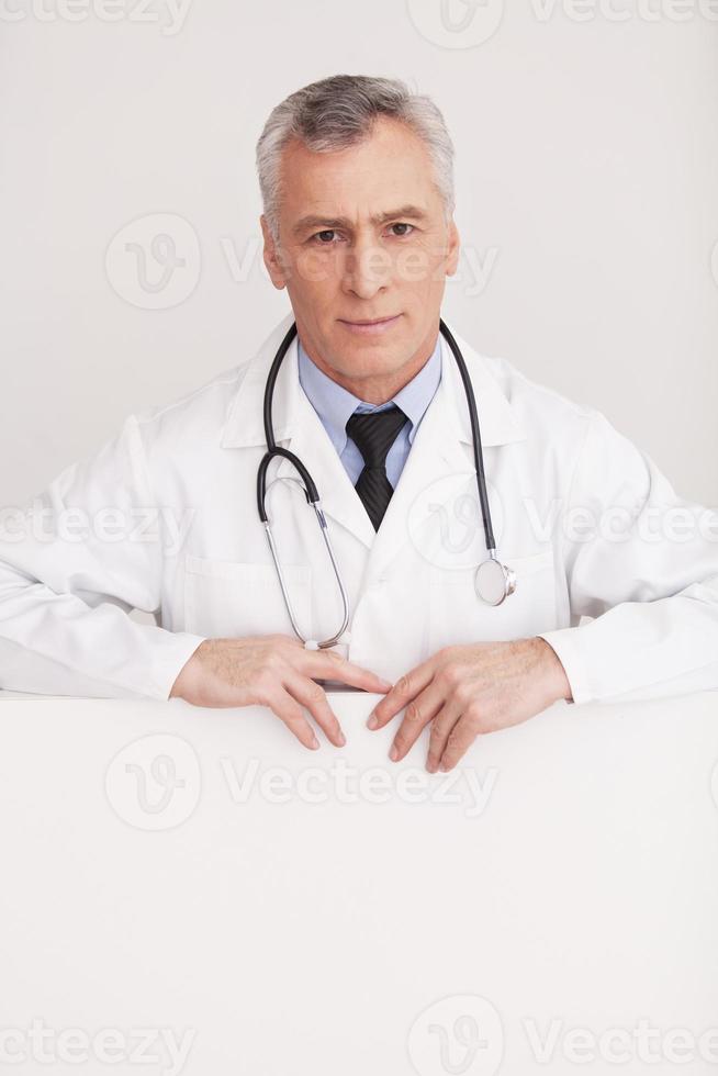 arzt lehnt sich an den kopierraum. leitender Arzt mit grauem Haar in Uniform, der aus dem Kopierbereich herausschaut und lächelt, während er isoliert auf Weiß steht foto
