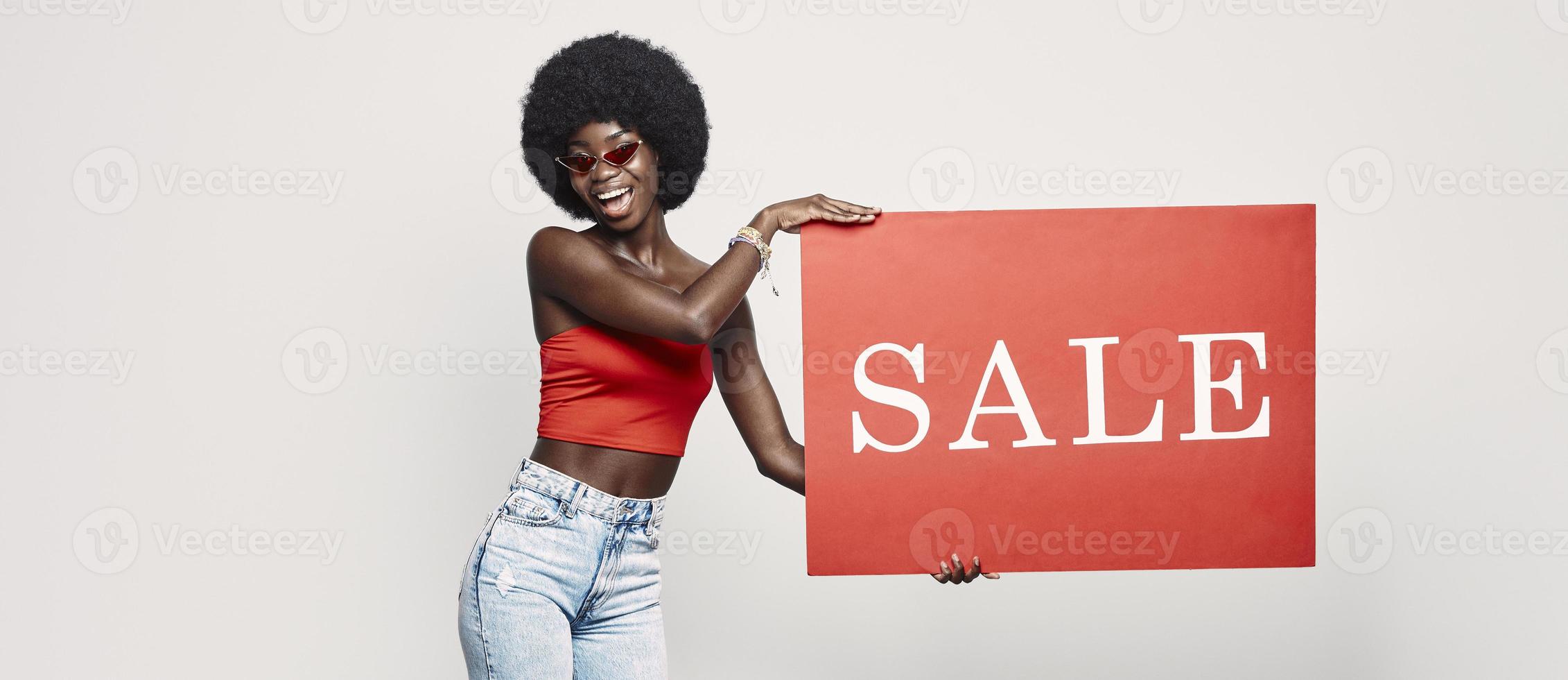 glückliche junge afrikanische frau, die verkaufsfahne hält und lächelt, während sie gegen grauen hintergrund steht foto