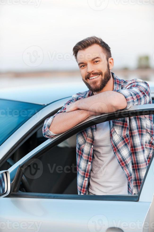 glücklicher Autobesitzer. glücklicher junger Mann, der sich an die Tür seines Autos lehnt und in die Kamera lächelt, während er im Freien steht foto