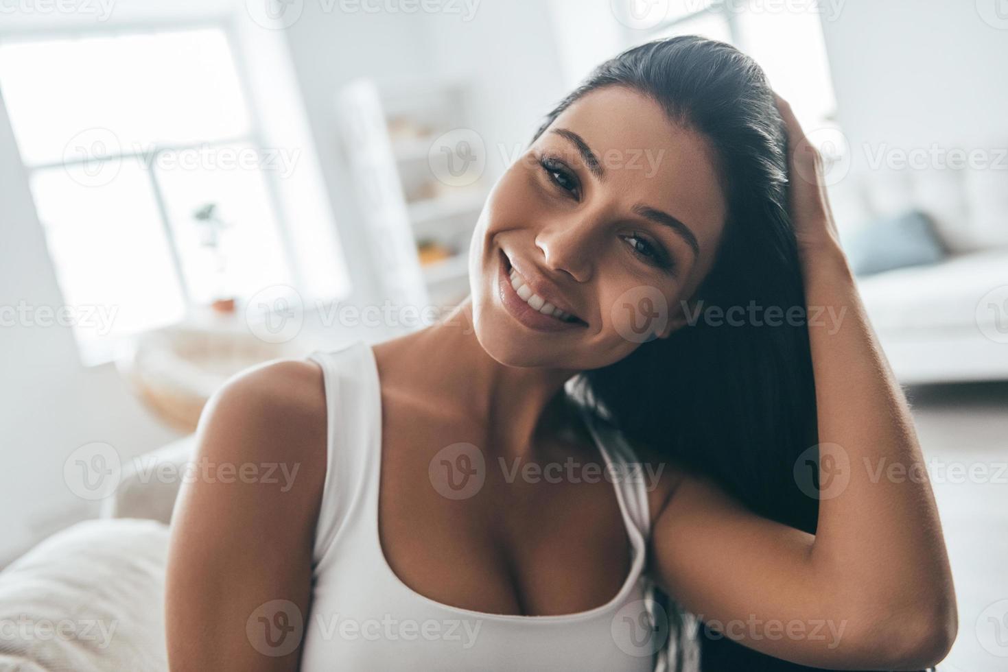Freizeit zu Hause genießen. Porträt einer attraktiven jungen Frau, die ihr Haar anpasst und in die Kamera schaut, während sie zu Hause auf der Couch sitzt foto