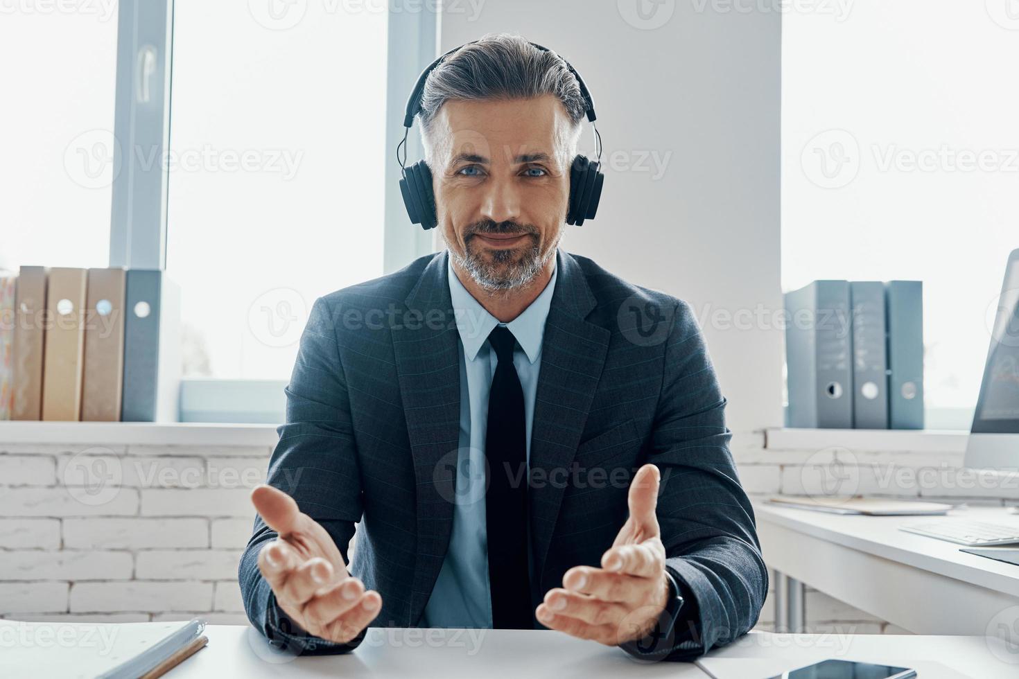 Selbstbewusster Geschäftsmann mit Kopfhörern, der lächelt und gestikuliert, während er an seinem Arbeitsplatz sitzt foto