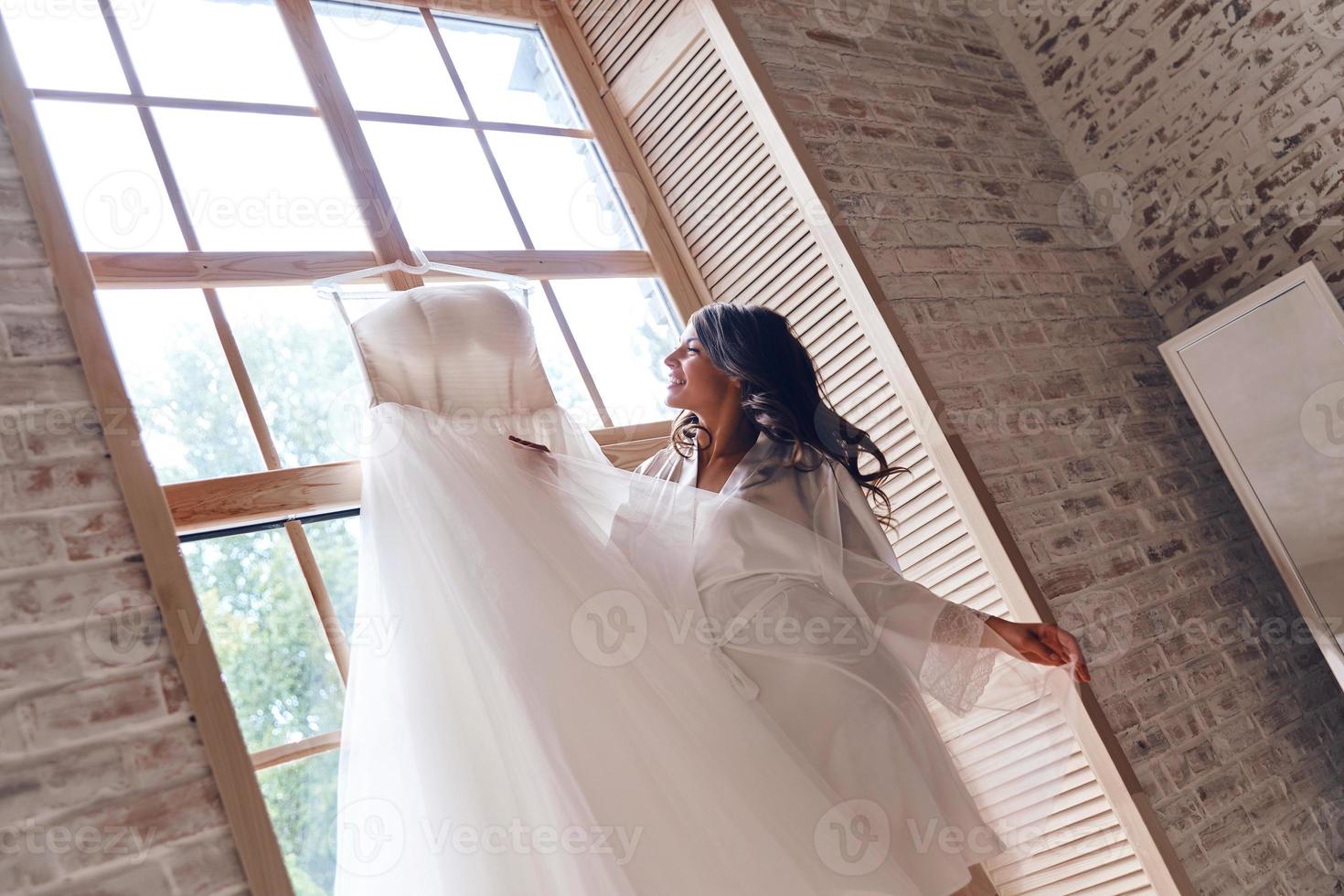 voller Freude. Schöne junge Frau im Seidenbademantel, die ihr Hochzeitskleid hält und lächelt, während sie in der Nähe des Fensters steht foto