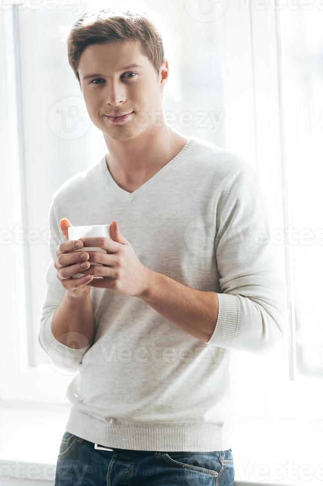 Morgenkaffee genießen. Fröhlicher junger Mann, der Kaffee trinkt und in die Kamera schaut, während er am Fenster steht foto