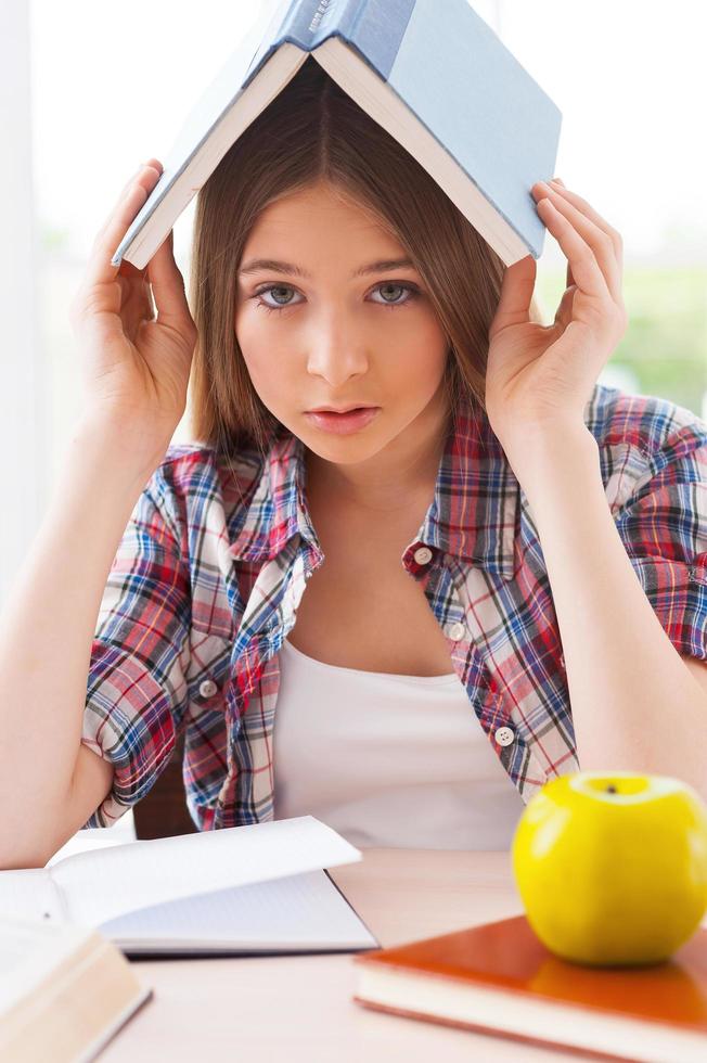 müde vom Lernen. frustriertes Teenager-Mädchen, das ein Buch auf dem Kopf trägt und in die Kamera schaut, während es am Schreibtisch sitzt und Bücher darauf liegen foto