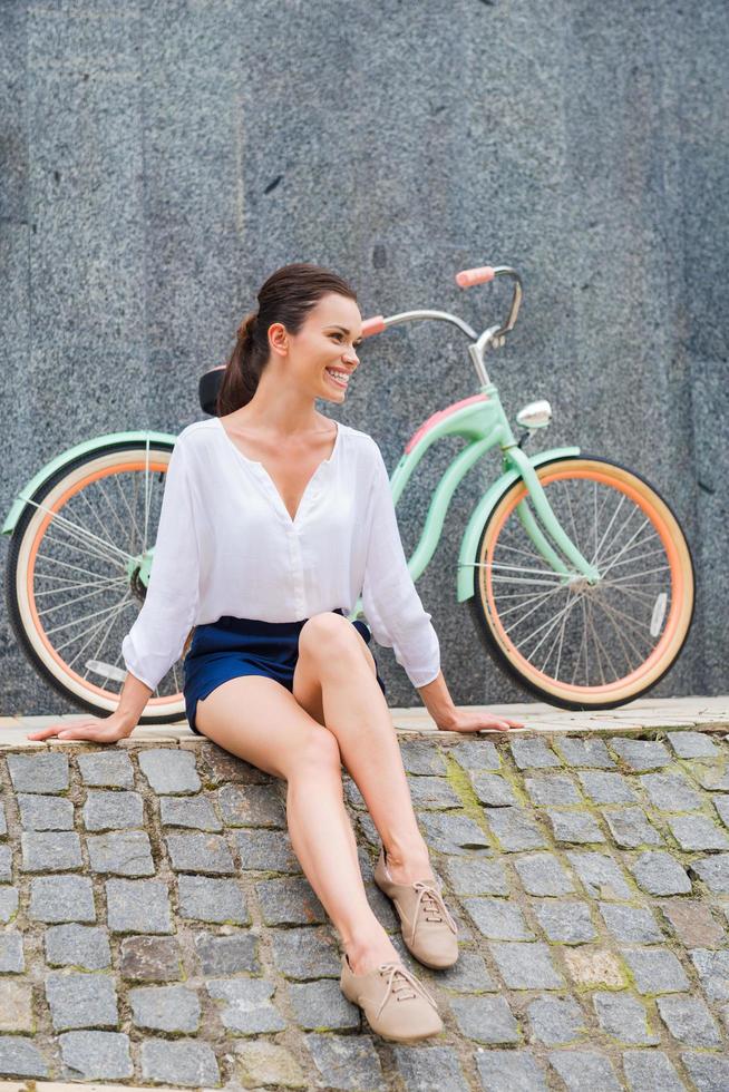 sorglos und stilvoll. Attraktive junge lächelnde Frau, die am Straßenrand und in der Nähe ihres Oldtimer-Fahrrads sitzt foto