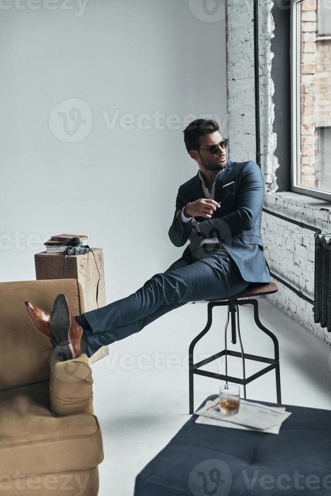 perfekter Stil. charmanter junger Mann im vollen Anzug, der wegschaut und die Arme verschränkt hält, während er auf dem Hocker sitzt foto