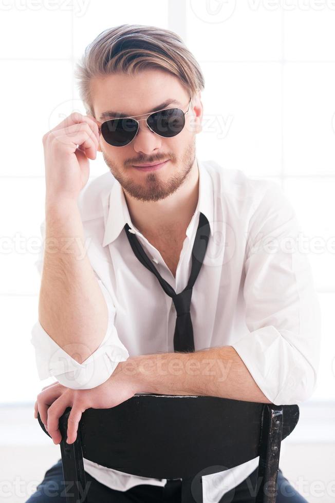 flirty fühlen. nachdenklicher junger Mann, der seine Sonnenbrille anpasst und in die Kamera schaut, während er auf dem Stuhl sitzt foto