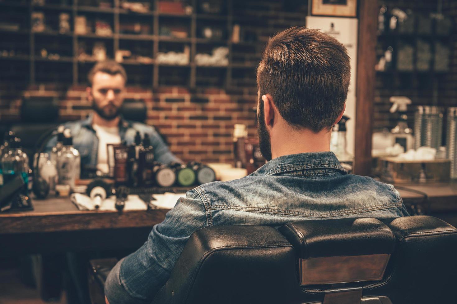 Zeit für schnelles Trimmen. Rückansicht eines hübschen jungen bärtigen Mannes, der sein Spiegelbild im Spiegel betrachtet, während er im Friseursalon auf einem Stuhl sitzt foto