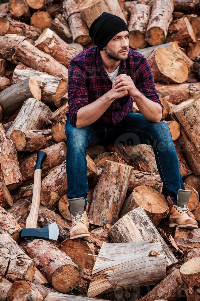 nach schwerer körperlicher Arbeit. voller Länge des nachdenklichen jungen Försters, der die Hände gefaltet hält und wegschaut, während er auf Baumstämmen sitzt foto