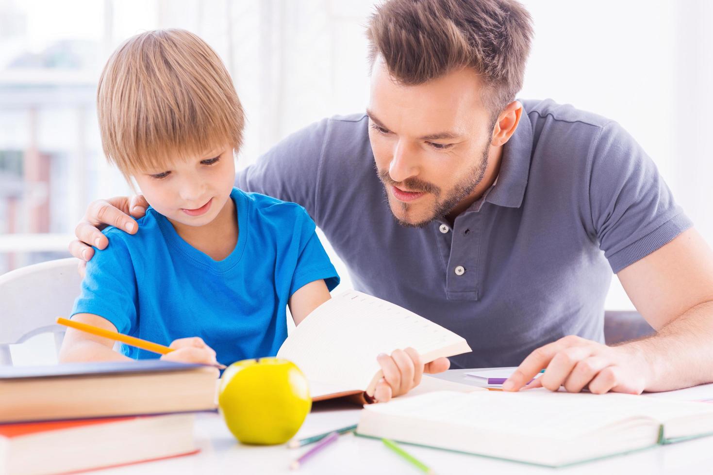 Sohn bei den Hausaufgaben helfen. Selbstbewusster junger Vater hilft seinem Sohn bei den Hausaufgaben, während er zusammen am Tisch sitzt foto