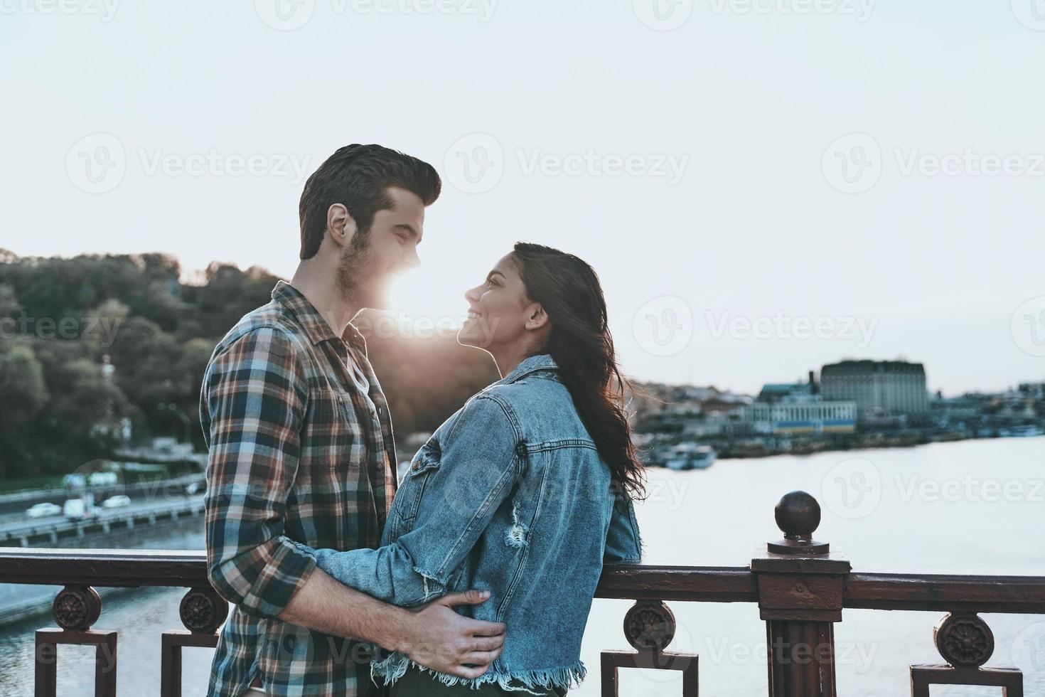Die Zeit bleibt stehen, wenn sie in der Nähe ist. Schönes junges Paar, das sich umarmt und ansieht, während es draußen auf der Brücke steht foto