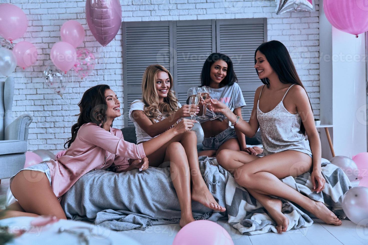 jubelt uns zu, vier attraktive junge lächelnde Frauen in Pyjamas, die einander anstoßen, während sie im Schlafzimmer eine Pyjamaparty mit Luftballons im ganzen Raum veranstalten foto