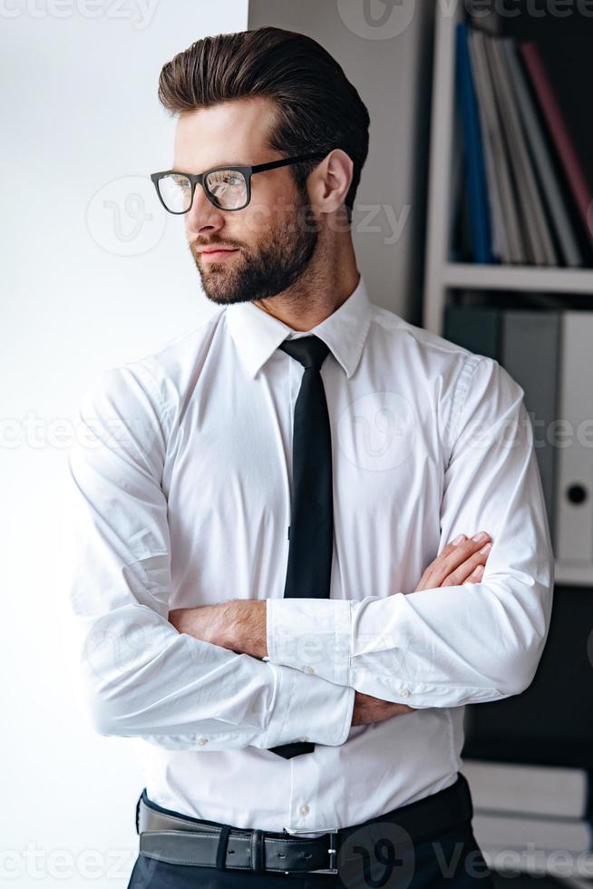 Suche nach neuer Lösung. seriöser junger Geschäftsmann mit Brille, der die Arme verschränkt hält und wegschaut, während er im Büro steht foto