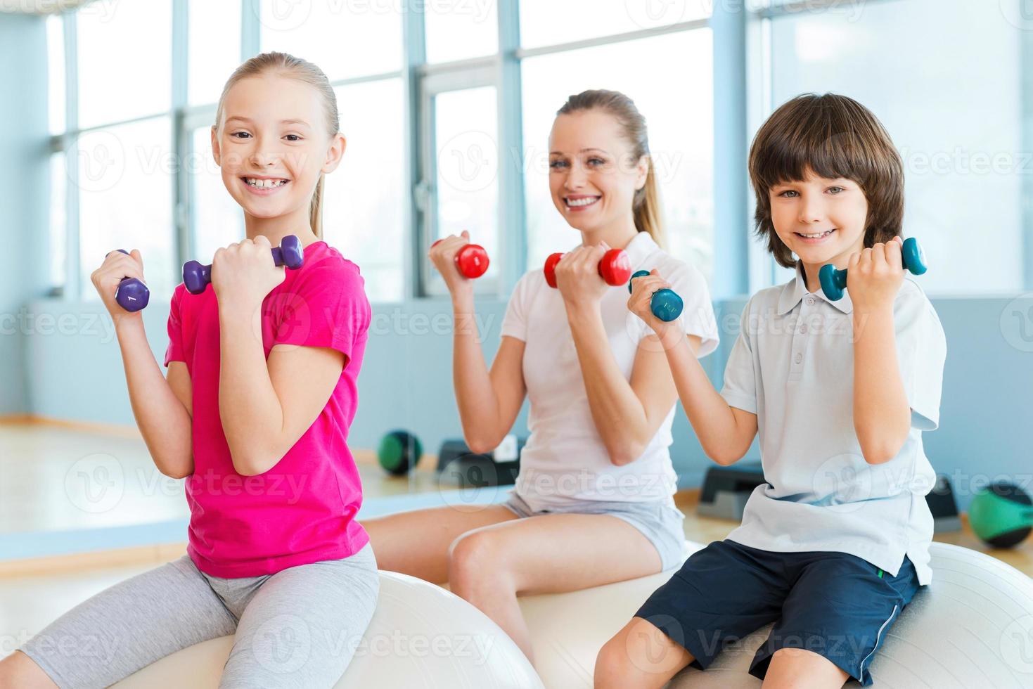 unseren Körper fit zu halten. Fröhliche Mutter und zwei Kinder trainieren mit Hanteln im Fitnessstudio, während sie zusammen auf den Fitnessbällen sitzen foto