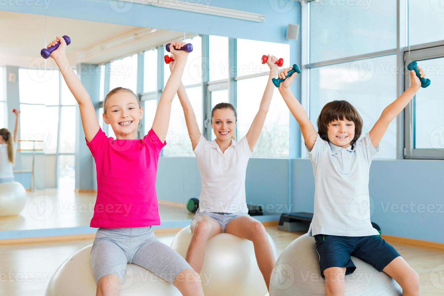 genießen Sie einen gesunden Lebensstil. Fröhliche Mutter und zwei Kinder trainieren mit Hanteln im Fitnessstudio, während sie auf den Fitnessbällen sitzen foto